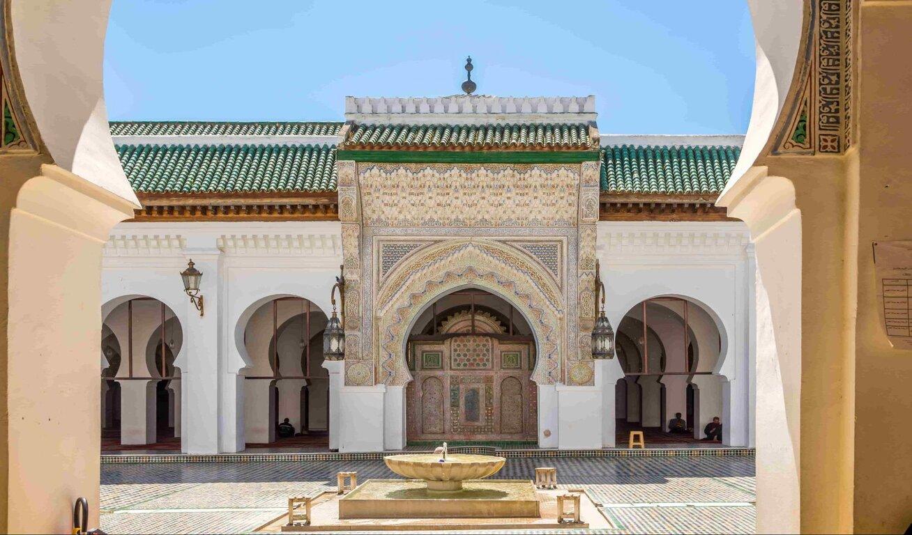 Exterior view of a mosque with a central fountain, green-tiled roof, and detailed archway, framed by two large pillars