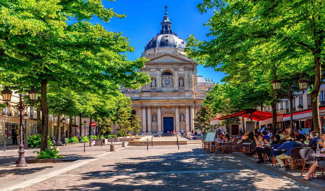 A street view with outdoor seating and trees leading to a historic building with a large dome and statues.