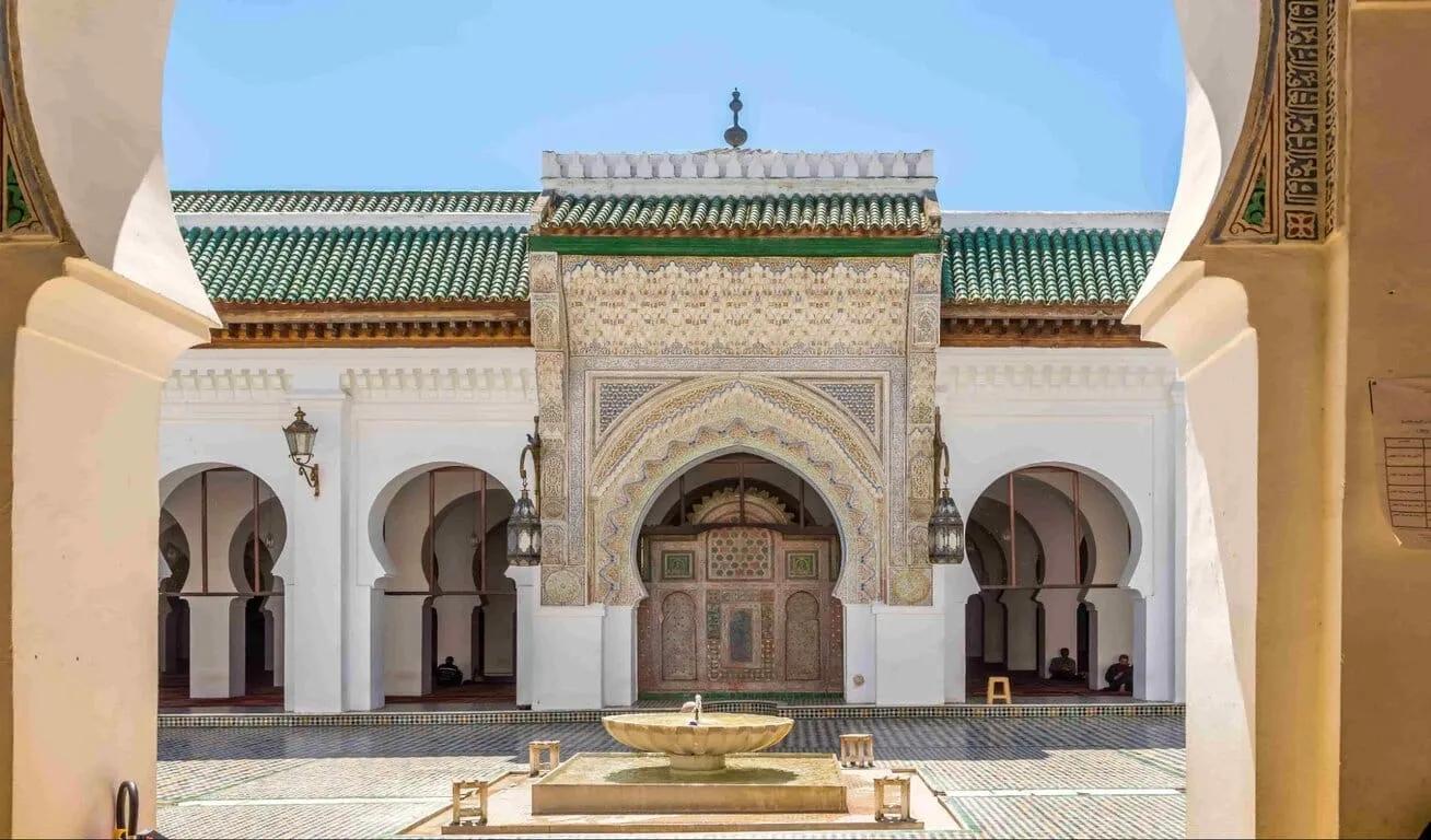 Exterior view of a mosque with a central fountain, green-tiled roof, and detailed archway, framed by two large pillars