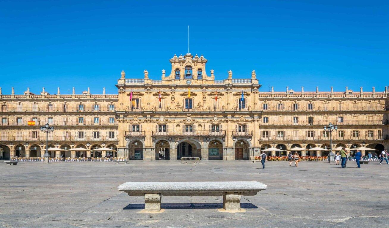 Wide shot of a historic building in a large plaza, with intricate stonework and multiple flags, under a clear blue sky.
