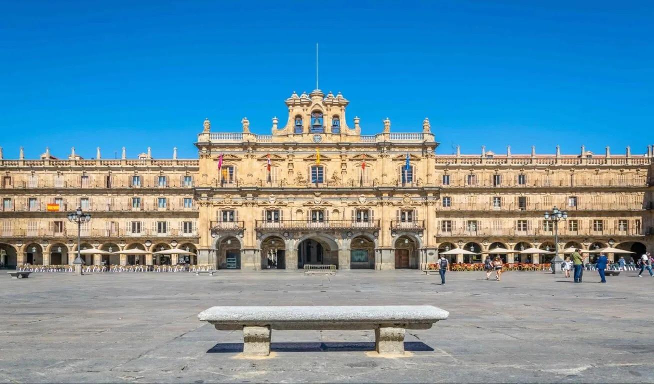 Wide shot of a historic building in a large plaza, with intricate stonework and multiple flags, under a clear blue sky.